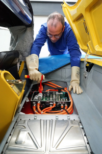 Unter dem Verdeck-Raum einer gelben Corvette C1 sieht man elektronische Komponenten. Ein Ladegerät sowie eine Hochvolt-Stromverteilerbox. Die Stromverteilerbox ist offen und wird gerade von einer Person mit Schutzhandschuhen angeschlossen.