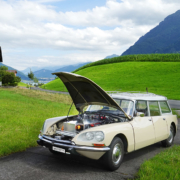 Beige Citroen DS Break steht mit offener Motorhaube auf einem Einfahrtssträsschen im Grünen. Statt des Motors sieht man eine Batteriekiste. Im Hintergrund die Rigi.