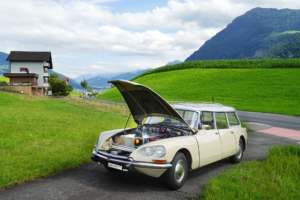 Beige Citroen DS Break steht mit offener Motorhaube auf einem Einfahrtssträsschen im Grünen. Statt des Motors sieht man eine Batteriekiste. Im Hintergrund die Rigi.