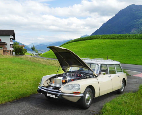 Beige Citroen DS Break steht mit offener Motorhaube auf einem Einfahrtssträsschen im Grünen. Statt des Motors sieht man eine Batteriekiste. Im Hintergrund die Rigi.