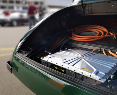 Das Heck eines grünen Jaguar XK 150, ohne Kofferraumklappe. Man sieht ein Ladegerät und orange Kabel.