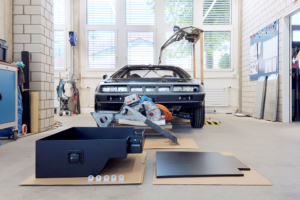 Im Vordergrund die Batteriekiste und deren Deckel. Dahinter die Motorenhalterung und der Motor. Im Hintergrund das Fahrzeug von vorne, eine Flügeltüre geöffnet.