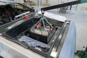 Der Frontraum des Fahrzeugs, die Batteriekiste wird gerade hineingestellt. Sie hängt an einem mobilen Kran und ist mit Ketten daran befestigt.