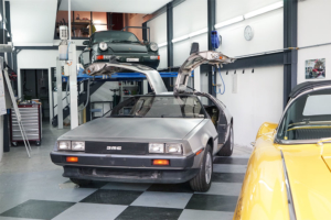 DeLorean DMC-12 in einer Werkstatt. Von vorne, leicht seitlich. Die Flügeltüren sind offen. Im Hintergrund ein Porsche 911 auf einem Fahrzeuglift. Im Vordergrund ein Teil einer gelben Corvette C1.