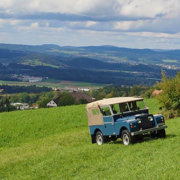 Ein blauer Landrover Series I mit beigem Verdeck fährt in einer hügeligen Landschaft eine Wiese hoch.