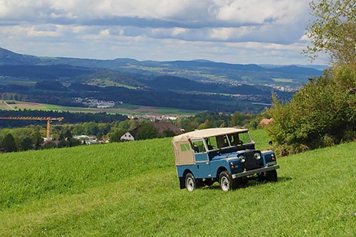 Ein blauer Landrover Series I mit beigem Verdeck fährt in einer hügeligen Landschaft eine Wiese hoch.