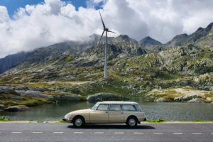 Beige Citroen DS Break auf der Gotthard-Passhöhe. Dainter Berglandschaft und ein Windrad.