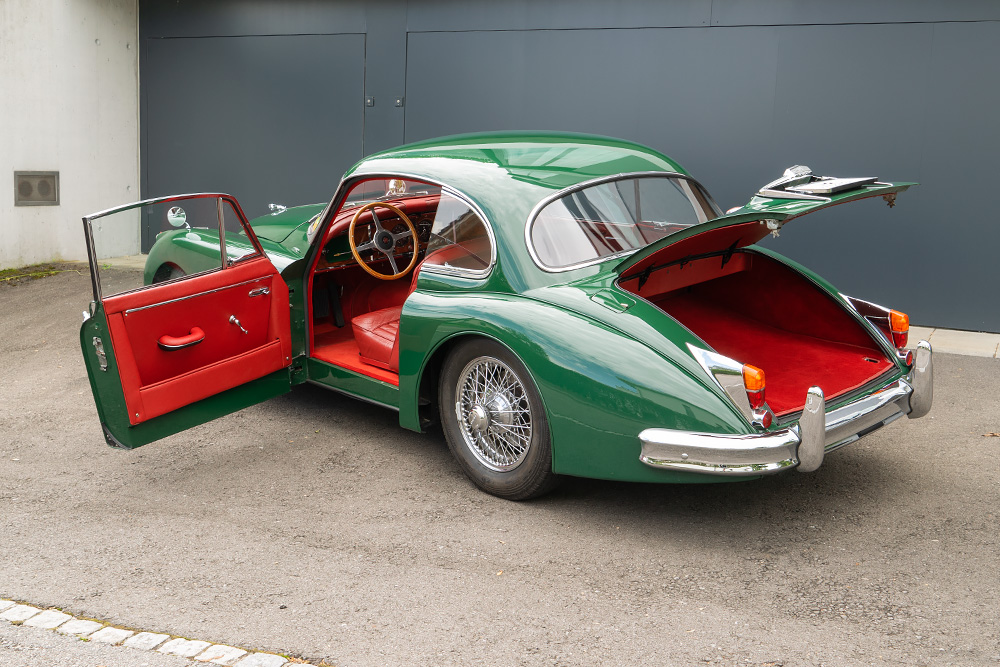 Ein grüner Jaguar XK150 mit rotem Interieur steht mit offenen Türen und Kofferraum vor einer Garage.