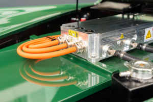 Blick in den Motorenraum des Jaguars. Man sieht den Deckel der Batteriekiste, in Wagenfarbe lackiert (grün), sowie den Umrichter, von welchem aus drei dicke, orange Hochvolt-Kabel im Bogen nach hinten gezogen werden.