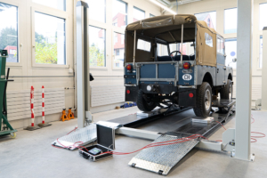 Ein Land Rover Series I in blau auf einem Viersäulenlift, aufgenommen von schräg hinten. Seine Räder stehen auf Waagen.