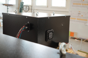 Blick auf die vordere Batteriekiste des eLandys, vor deren Einbau. Die Kiste ist schwarz. Man sieht den Niedervolt-Stecker mit diversen Kabeln, sowie ein Ventil zur Entgasung.
