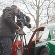 Ein Kameramann von hinten. Er steht neben einem Jaguar XK 150 mit offener Türe, und filmt durch diese das Interieur des Fahrzeugs. Im Hintergrund leicht vom Nebel verhüllte spätwinterliche Bäume.