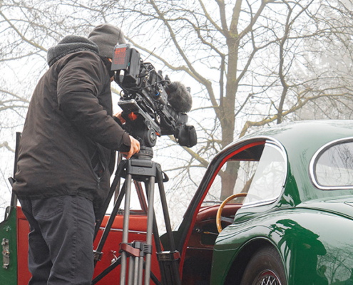Ein Kameramann von hinten. Er steht neben einem Jaguar XK 150 mit offener Türe, und filmt durch diese das Interieur des Fahrzeugs. Im Hintergrund leicht vom Nebel verhüllte spätwinterliche Bäume.