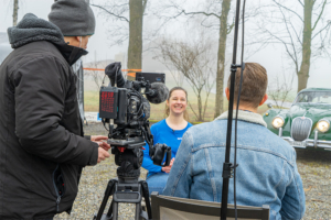 Links im Bild ein Kameramann mit Kamera auf dem Stativ, von hinten aufgenommen. Rechts ein Reporter auf, sitzend auf einem Stuhl, ebenfalls von hinten. Zwischen Kamera und Reporter sieht man Silvia Maron während des TV-Interviews. Im Hintergrund rechts der Jaguar XK 150
