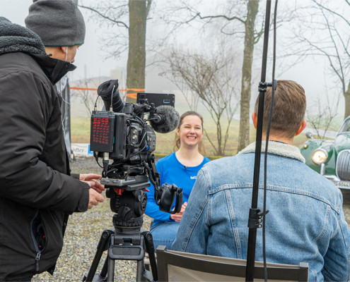 Links im Bild ein Kameramann mit Kamera auf dem Stativ, von hinten aufgenommen. Rechts ein Reporter auf, sitzend auf einem Stuhl, ebenfalls von hinten. Zwischen Kamera und Reporter sieht man Silvia Maron während des TV-Interviews. Im Hintergrund rechts der Jaguar XK 150