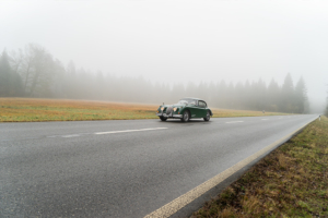 Ein Jaguar XK 150 in grün fährt auf einer Landstrasse durch den Nebel. Im Hintergrund erkennt man Wiese und Wald.