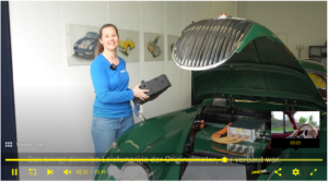 Ausschnitt aus einem Video. Ein Jaguar XK 150 mit offener Fronthaube, im ehemaligen Motorenraum sieht man Batterie und Umrichter. Daneben Silvia Marton in blauem Shirt, in der Hand ein Dummy des im Fahrzeug verbauten Elektromotors.