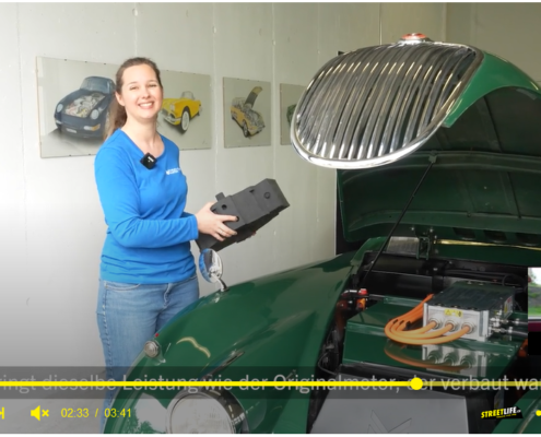 Ausschnitt aus einem Video. Ein Jaguar XK 150 mit offener Fronthaube, im ehemaligen Motorenraum sieht man Batterie und Umrichter. Daneben Silvia Marton in blauem Shirt, in der Hand ein Dummy des im Fahrzeug verbauten Elektromotors.