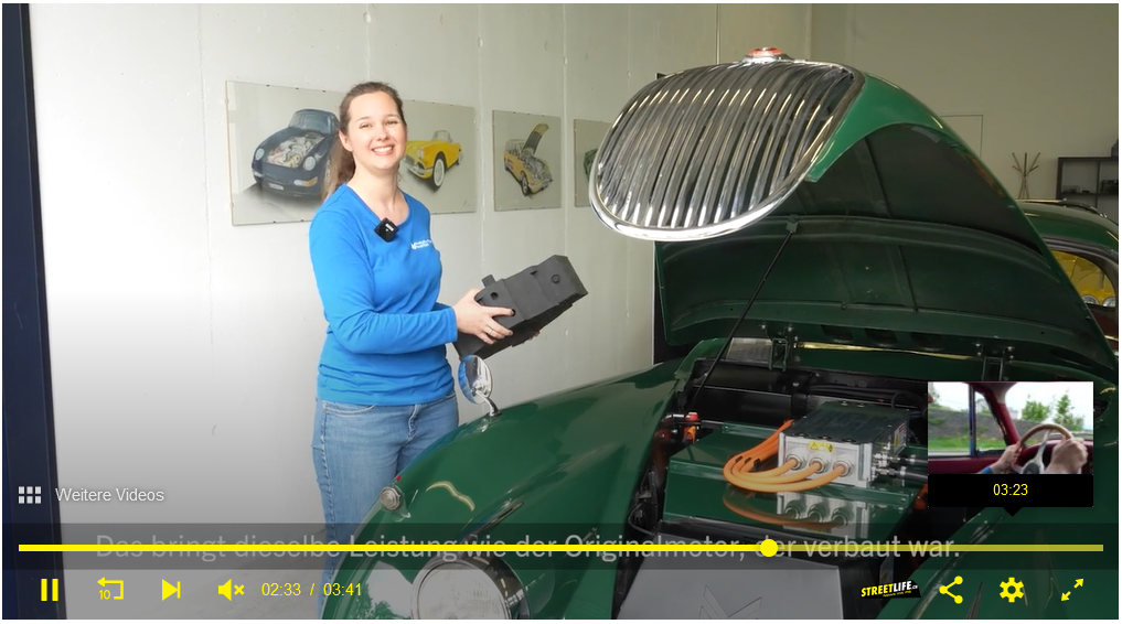 Ausschnitt aus einem Video. Ein Jaguar XK 150 mit offener Fronthaube, im ehemaligen Motorenraum sieht man Batterie und Umrichter. Daneben Silvia Marton in blauem Shirt, in der Hand ein Dummy des im Fahrzeug verbauten Elektromotors.