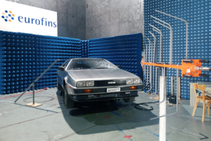 Ein DeLorean wird gerade auf seine elektromagnetische Verträglichkeit geprüft. Er steht in einer Prüfhalle, eine orange-graue Antenne ist auf ihn gerichtet. Antenne im Vordergrund, Fahrzeug im Hintergrund, von leicht schräg vorne fotografiert.