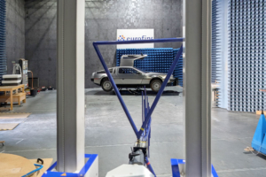Ein DeLorean wird gerade auf seine elektromagnetische Verträglichkeit geprüft. Er steht in einer Prüfhalle, eine blaue Antenne ist auf ihn gerichtet und misst seine elektromagnetischen Strahlungswerte. Man sieht das Fahrzeug im Hintergrund und die Antenne im Vordergrund.