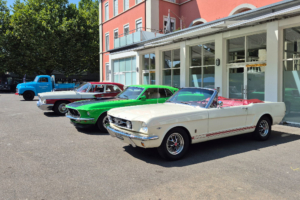 Nebeneinander stehende Fahrzeuge, seitlich aufgenommen. Zu vorderst ein weisser Mustang, Cabrio, dahinter ein gröner Mustang Mach I. Dahinter ein Chrysler Newport und ein Opel Blitz.