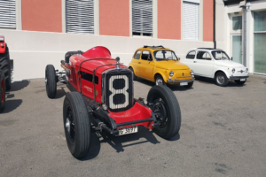 Links ein roter Nash Twin Ignition Eight in offener Bauweise. Sein Kühlergrill wird von einer 8 geziert. Rechts davon zwei Fiat Nuova 500, einer in gelb, einer in weiss.