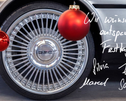 Bild eines Rads an einem Fahrzeug. Die Felge hat ein Speichen-Design, ein DMC-Logo verdeckt die Radmuttern. Vor dem Rad hängen zwei rote Christbaumkugeln ins Bild, eine davon matt, eine glänzend. Auf dem Bild steht zudem die hangeschriebene Nachricht "Wir wünschen frohe Festtage". Darunter die Unterschriften von Silvia, Till, Marcel und Sabrina.