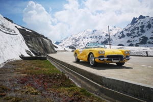 Eine gelbe Corvette ist am Rande einer Passstrasse parkiert. Links hat es einen Schneewall, im Hintergrund eine leicht verschneite Berglandschaft.