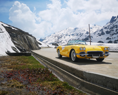 A yellow Corvette is parked on the side of a mountain road. There is a snow bank on the left and a lightly snow-covered mountain landscape in the background.