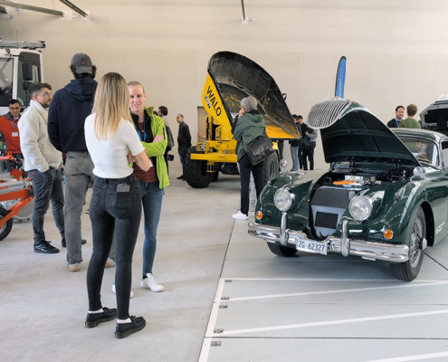 Verschiedene Fahrzeuge, unter anderem ein Muldenfahrzeug, ein Mäher sowie ein Jaguar XK 150 stehen in einem Hangar. Diverse Menschen betrachten und besprechen die Fahrzeuge
