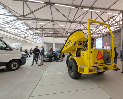 Verschiedene Fahrzeuge, unter anderem ein Muldenfahrzeug, ein Lieferwagen sowie ein Jaguar XK 150 stehen in einem Hangar. Diverse Menschen betrachten und besprechen die Fahrzeuge