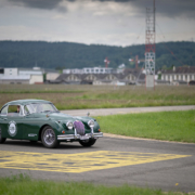 Ein grüner Jaguar XK 150 fährt über eine Flughafen-Fahrbahn. Gelbe Marierungen auf der Fahrbahn, im Hintergrund Baumarkierungen. Auf der Seite des Jaguars ist eine Beschriftung angebracht, die darauf hinweist, dass es sich um ein Elektroauto handelt.