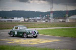 Ein grüner Jaguar XK 150 fährt über eine Flughafen-Fahrbahn. Gelbe Marierungen auf der Fahrbahn, im Hintergrund Baumarkierungen. Auf der Seite des Jaguars ist eine Beschriftung angebracht, die darauf hinweist, dass es sich um ein Elektroauto handelt.