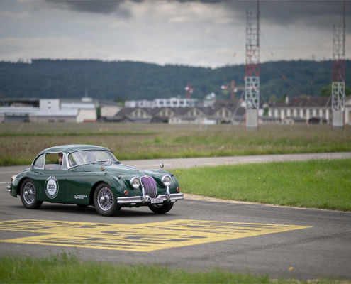 Ein grüner Jaguar XK 150 fährt über eine Flughafen-Fahrbahn. Gelbe Marierungen auf der Fahrbahn, im Hintergrund Baumarkierungen. Auf der Seite des Jaguars ist eine Beschriftung angebracht, die darauf hinweist, dass es sich um ein Elektroauto handelt.