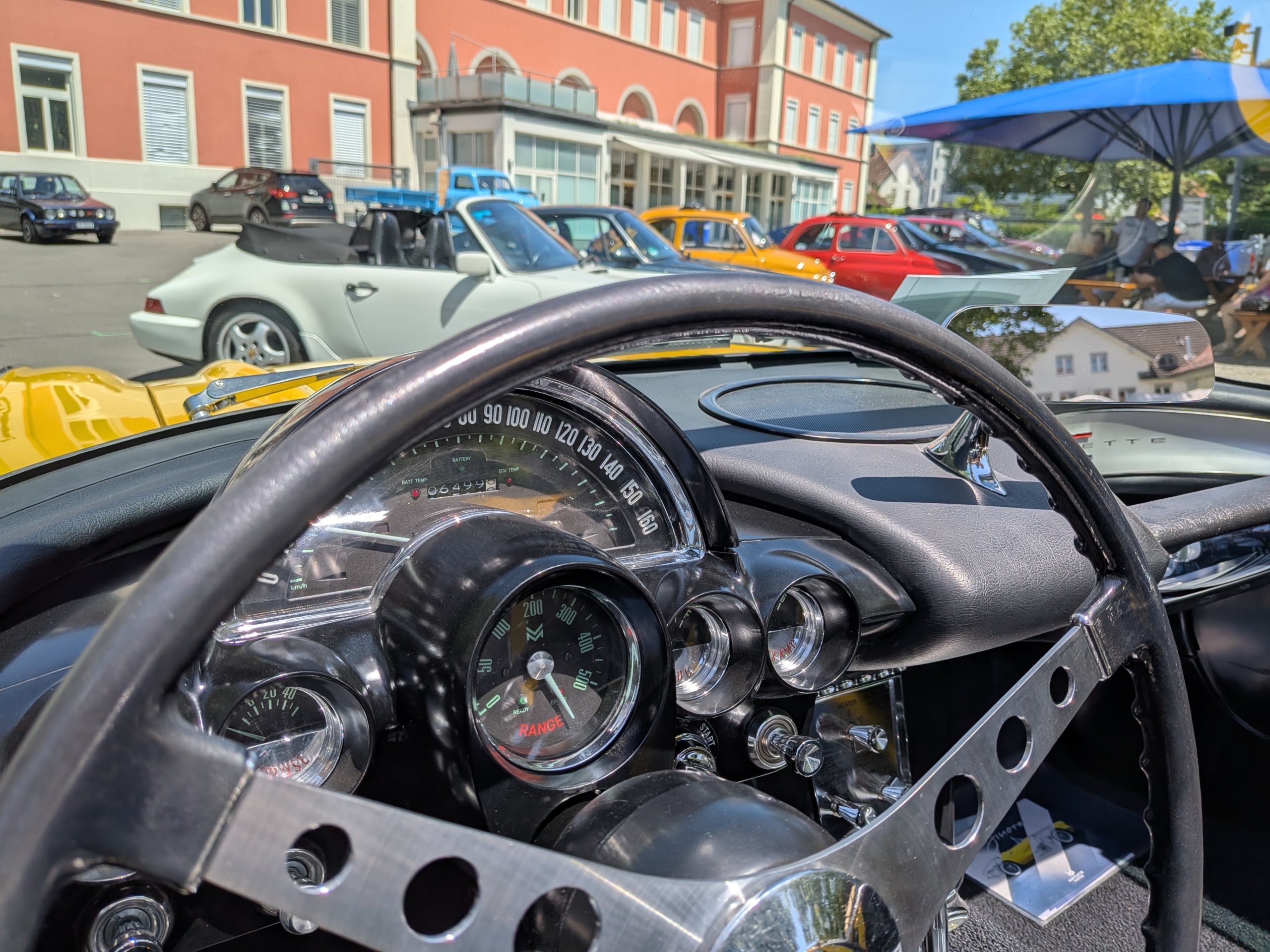 Blick über das Lenkrad einer Corvette C1. Die Armaturen verraten, dass es sich hier um ein Fahrzeug handelt, dass zum Elektroauto umgebaut wurde. Denn an Stelle des Tourenzählers zeigt die klassische Rundanzeige die Restreichweite an. Im Hintergrund sieht man diverse Fahrzeuge. Rechts eine Reihe mit diversen Fahrzeugen. Zwei Porsche 911 in weiss und grau, zwei Fiat Nuova 500 in gelb und rot sind gut erkennbar, dahinter lassen sich zwei Alfa Romeo GTV in rot und blau sowie eine Charleston-Ente erahnen.