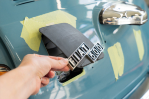 Ladeklappe, noch roh und schwarz, mit originalem Fiatschriftzug für den elektrifizierten Fiat 500. Eine Hand hält sie etwas offen.