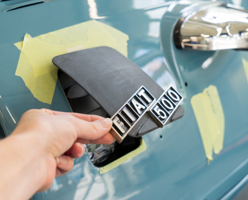 DSC09132_manufakturmartin_ladedose_blog Ladeklappe, noch roh und schwarz, mit originalem Fiatschriftzug für den elektrifizierten Fiat 500. Eine Hand hält sie etwas offen.