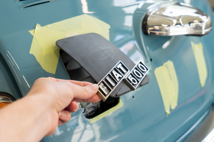 Ladeklappe, noch roh und schwarz, mit originalem Fiatschriftzug für den elektrifizierten Fiat 500. Eine Hand hält sie etwas offen.