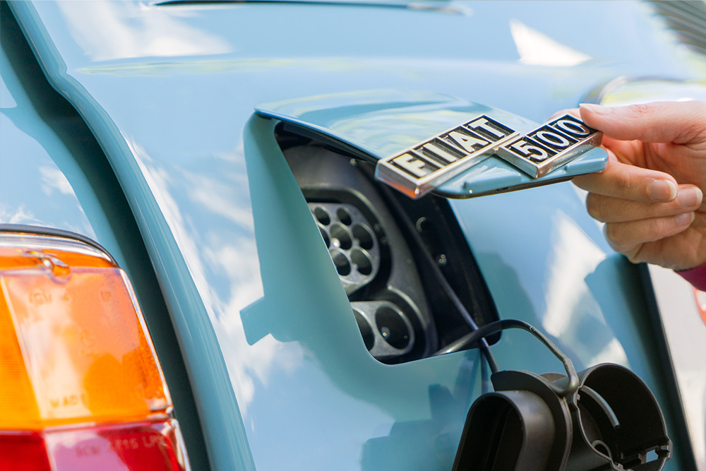 The finished charging flap An opening at the rear of the blue Fiat 500, which can be closed with a flap in the same colour as the car. In the picture, it is half-open and bears the “Fiat 500” logo.