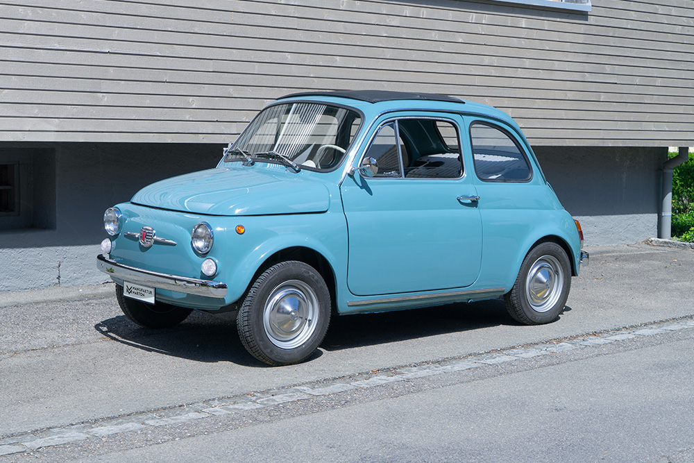 Fiat 500, newly restored and electrified A medium-blue Fiat Nuova 500 is parked in front of a building with a wooden façade. The car is photographed from the side.