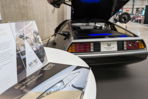 Ein runder Bartisch im Vordergrund, auf dem Flyer eines Autofolierers liegen. Dahinter sichtbar das Heck eines DeLorean.
