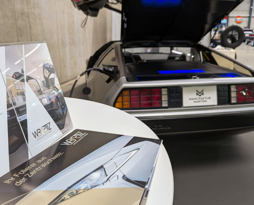manufakturmarton_SCW2026_20260529_090017196 Ein runder Bartisch im Vordergrund, auf dem Flyer eines Autofolierers liegen. Dahinter sichtbar das Heck eines DeLorean.