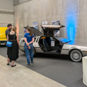 Messehalle mit Sichtbetonwand. Zwei Fahrzeuge, ein DeLorean sowie eine gelbe Corvette sind austestellt. Einige Leute stehen im Bild und sprechen.