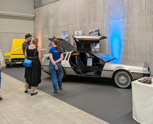 manufakturmarton_SCW2026_20260529_124806887 Messehalle mit Sichtbetonwand. Zwei Fahrzeuge, ein DeLorean sowie eine gelbe Corvette sind austestellt. Einige Leute stehen im Bild und sprechen.