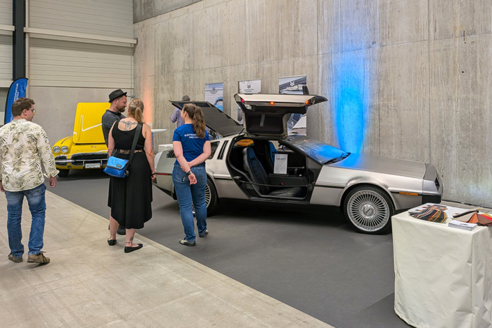 manufakturmarton_SCW2026_20260529_124806887 Messehalle mit Sichtbetonwand. Zwei Fahrzeuge, ein DeLorean sowie eine gelbe Corvette sind austestellt. Einige Leute stehen im Bild und sprechen.