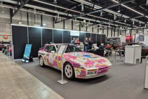 Ein Porsche 944 mit Rallye-Bemahlung an einem Messestand. Die Bemahlung ist Pink und Weiss.
