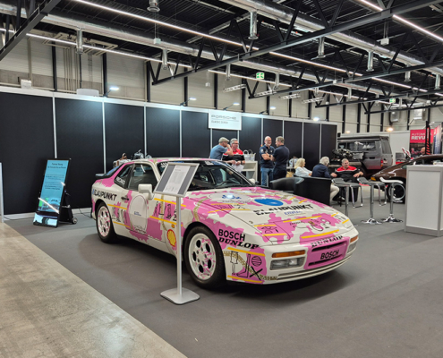manufakturmarton_SCW2026_20260530_173634 Ein Porsche 944 mit Rallye-Bemahlung an einem Messestand. Die Bemahlung ist Pink und Weiss.